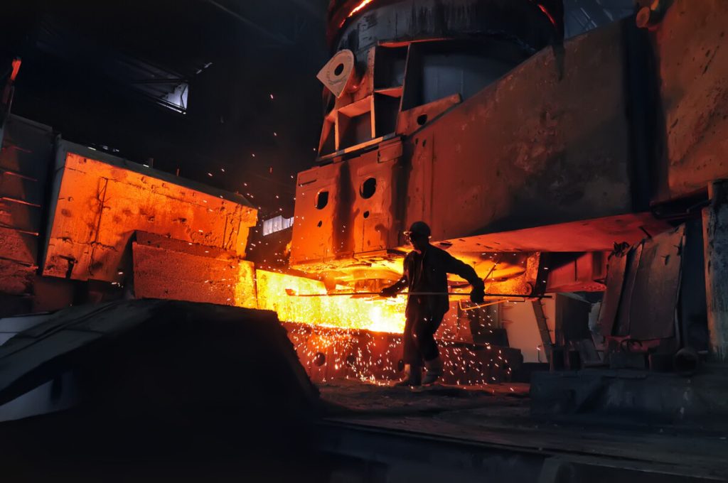 A worker stands near molten metal and flying sparks inside a brightly lit industrial foundry, where improved safety management helps protect employees in hazardous environments.