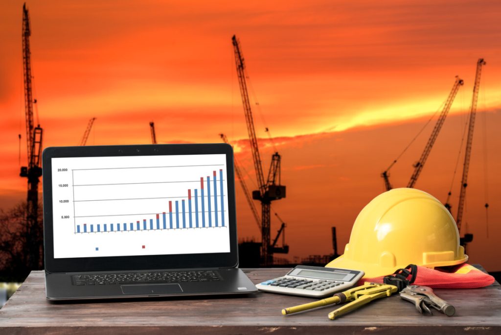 Laptop with bar chart, calculator, hard hat, and tools on table; cranes and sunset sky in background—illustrating digitizing document control in modern construction projects.