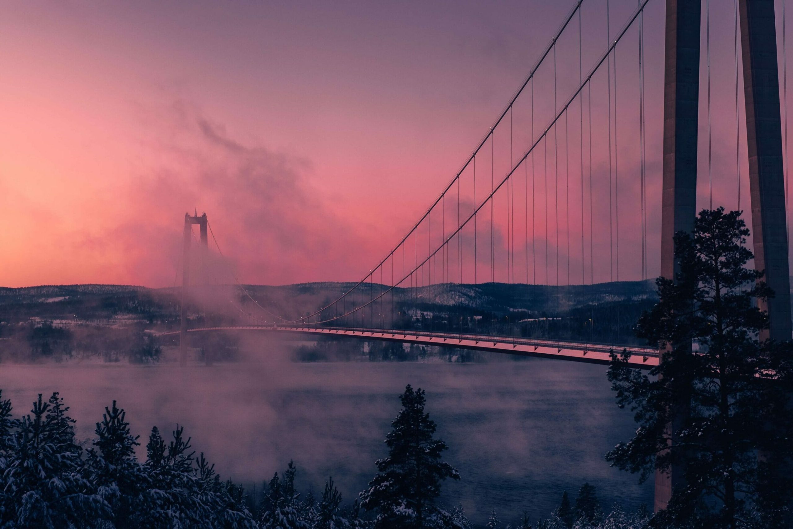 A suspension bridge spans a misty river at sunset, with snow-covered trees and hills in the background—a serene scene that subtly reflects the harmony sought in corporate sustainability efforts.