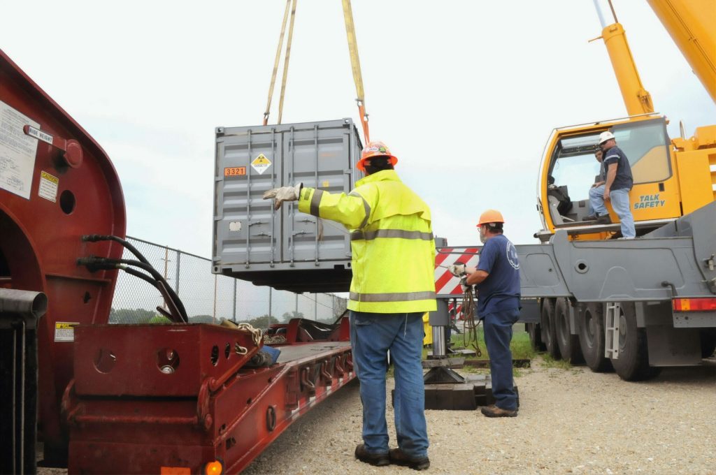 Workers guide a shipping container being lifted by a crane onto a trailer at an outdoor site, staying alert to human factor safety risks throughout the operation.