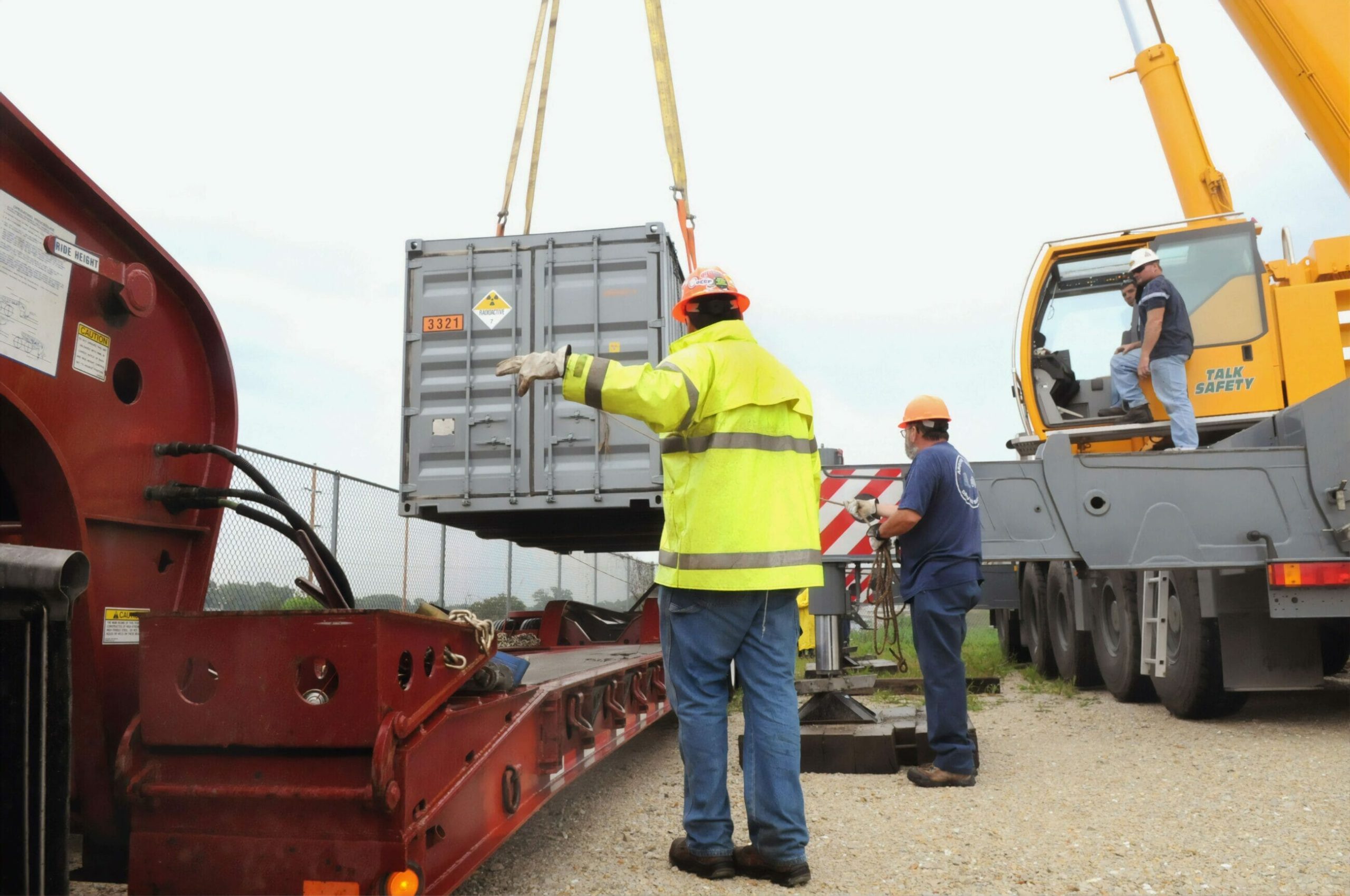 Workers guide a shipping container being lifted by a crane onto a trailer at an outdoor site, staying alert to human factor safety risks throughout the operation.