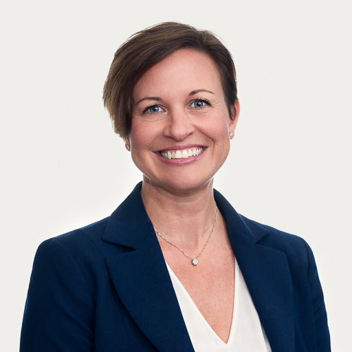 Smiling woman with short brown hair wearing a navy blazer over a white top, and a delicate necklace, posing against a plain white background.