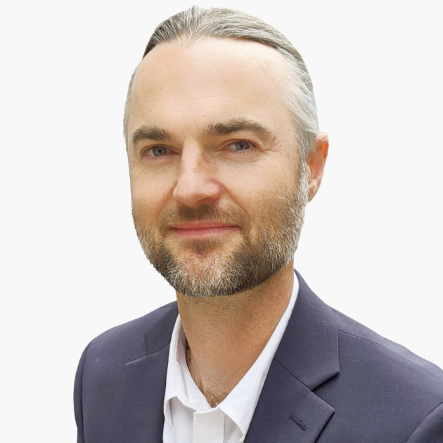 A man with light skin, blue eyes, and brown hair pulled back, wearing a navy blazer and white shirt, smiles slightly against a plain white background.