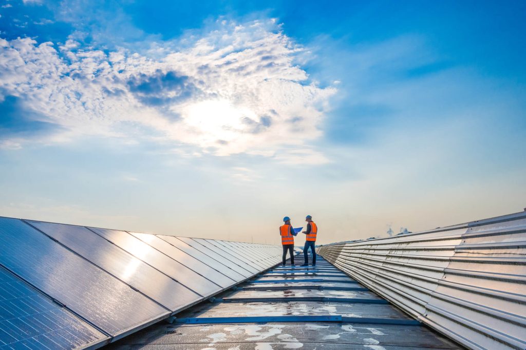 Two workers in orange vests and helmets stand on a rooftop next to solar panels, talking under a bright blue sky with scattered clouds and sunlight.