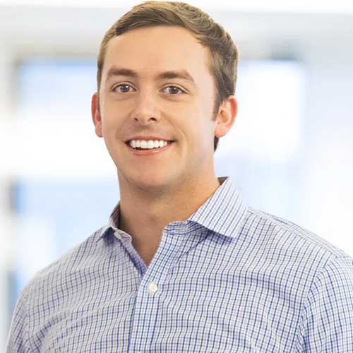 A young man with short brown hair smiles while wearing a light blue, checkered button-up shirt. The background is softly blurred with natural light.
