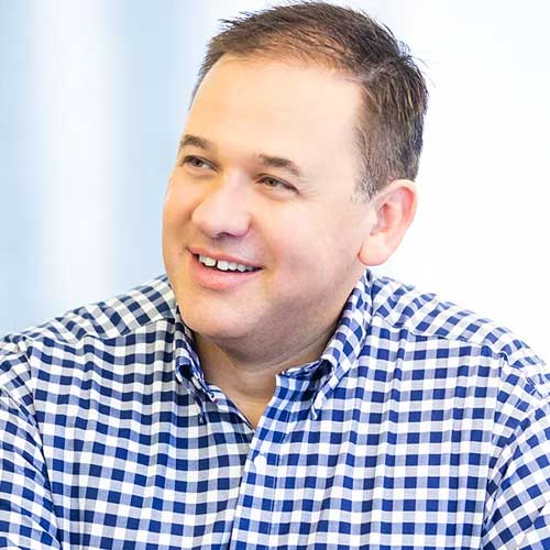 A smiling man with short brown hair wearing a blue and white checkered shirt, sitting and looking slightly to the left against a light background.