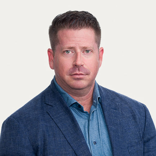 A man with short brown hair wearing a blue blazer and light blue collared shirt, looking directly at the camera with a neutral expression against a plain background.