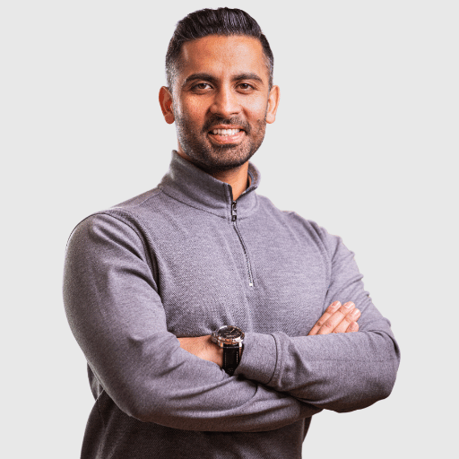 A man with short dark hair and a beard is standing with arms crossed, smiling. He is wearing a gray quarter-zip sweater and a watch, set against a light gray background.