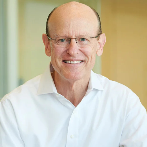 A smiling older man with glasses, a bald head, and a white collared shirt, standing indoors with a blurred background.