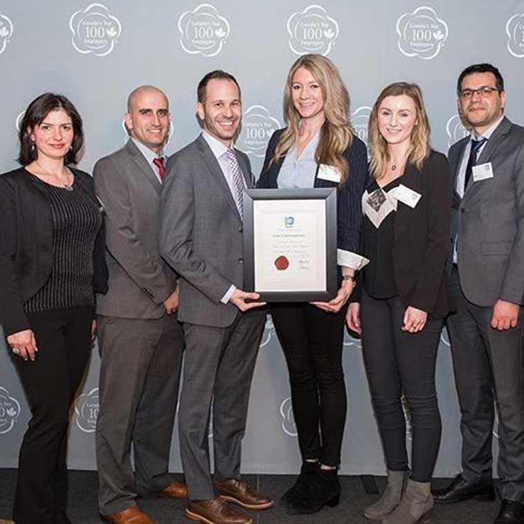 Six professionally dressed people stand in a row, smiling at the camera. The person in the middle holds a framed certificate, celebrating success. Behind them is a grey backdrop with white award logos, reflecting the achievements of careers at Cority.