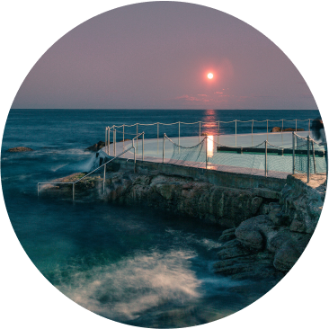 A round image of an ocean pool at dusk, with calm waves, a stone edge, and rope barriers. The full moon glows above the horizon, casting light on the water and evoking serenity—an ambiance echoing the calm progress of sustainability at Cority.