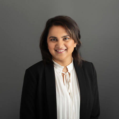A woman with dark hair wearing a black blazer and a white blouse smiles in front of a plain gray background.