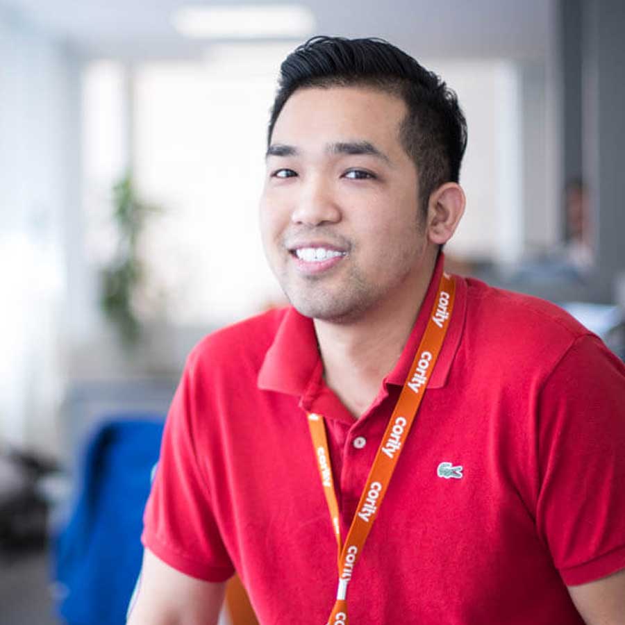 A man wearing a red polo shirt and an orange lanyard smiles at the camera in a bright office setting, reflecting the friendly atmosphere of Careers at Cority.
