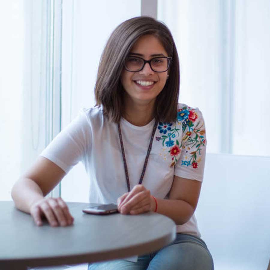 A young woman with shoulder-length brown hair and glasses sits at a table, smiling. She wears a white t-shirt with colorful floral embroidery on the shoulder and has a smartphone in front of her, exploring Careers at Cority.