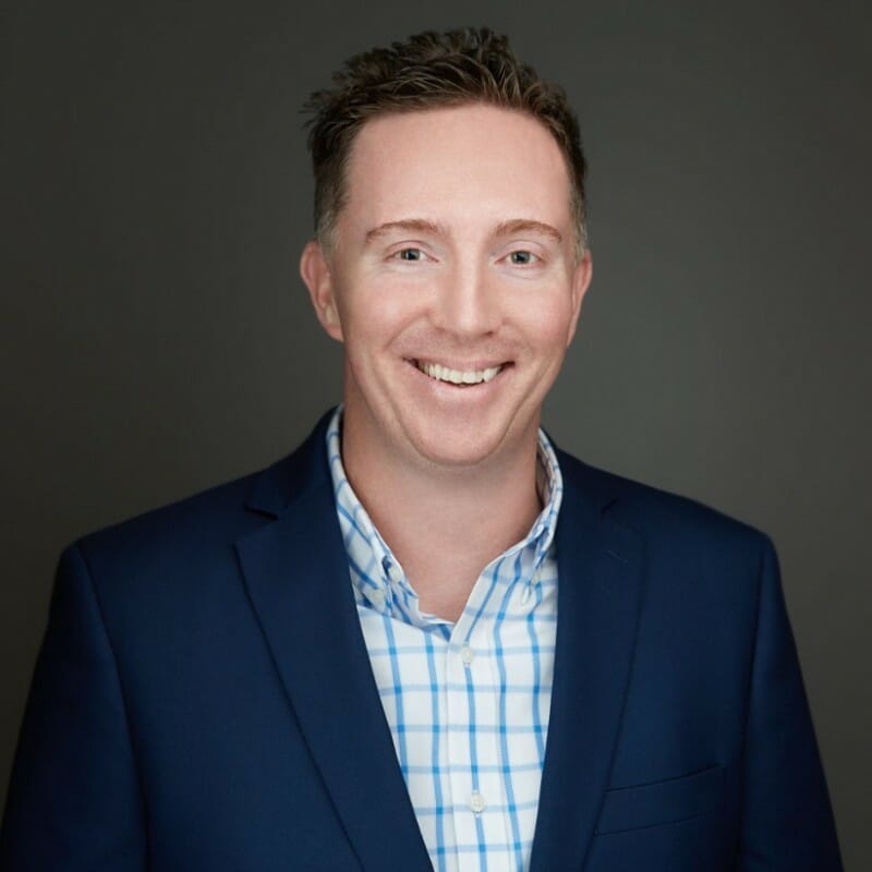 A man with short brown hair wearing a navy blue blazer and a blue-and-white checkered shirt smiles in front of a plain dark gray background.