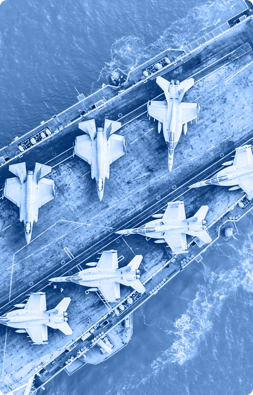 Aerial view of six fighter jets arranged on the deck of an aircraft carrier at sea, with waves visible around the ship. The image has a blue color filter, suitable for use in an Industry Tab Section.