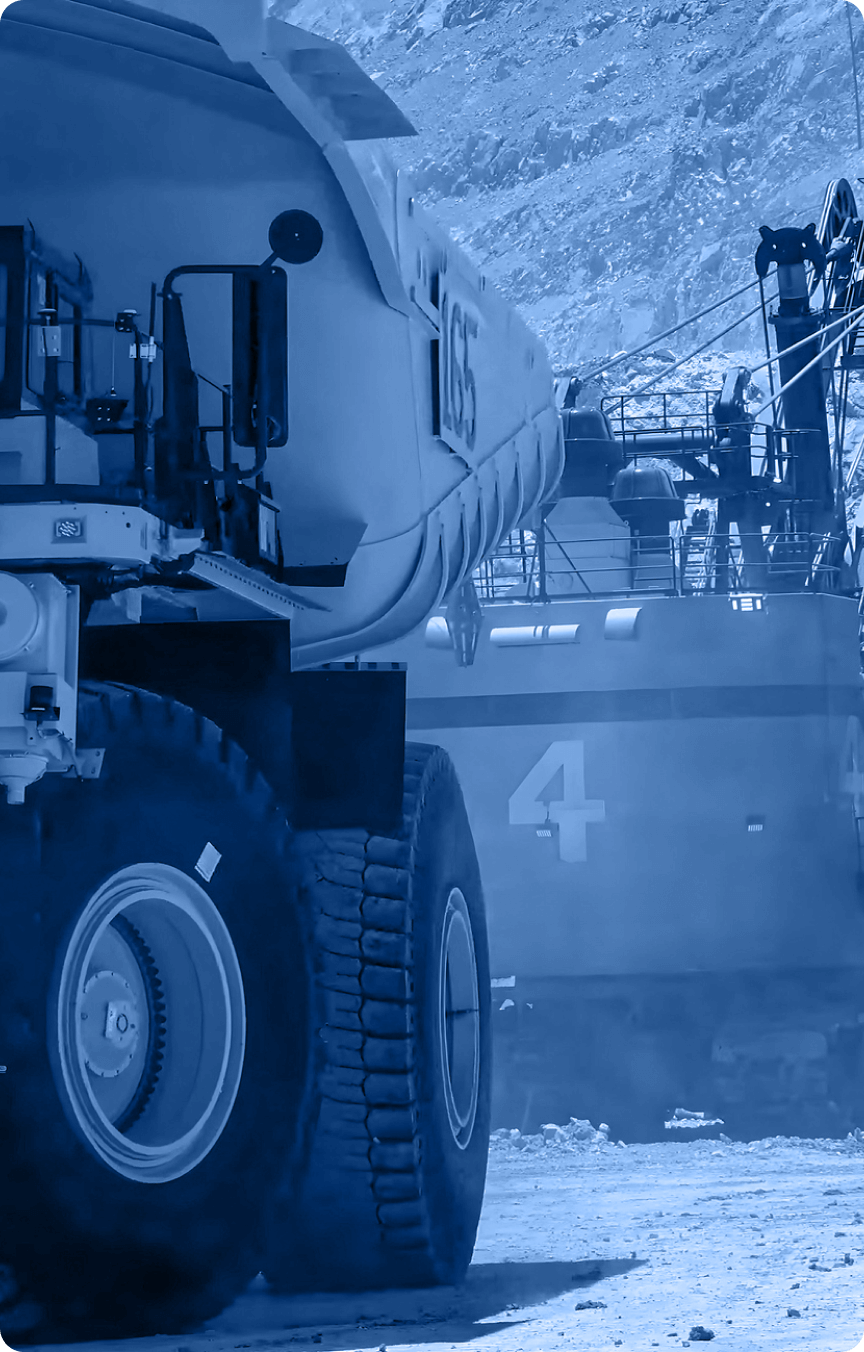 A large mining truck with oversized wheels is parked in front of a power shovel at an open-pit mining site, featured in the Industry Tab Section, with rocky terrain in the background. The image has a blue color tone.