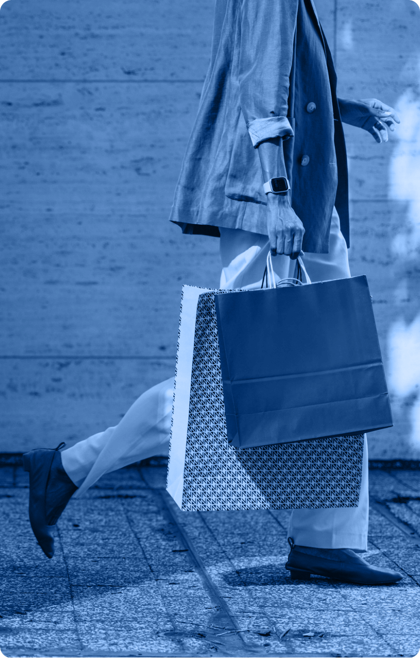 A person wearing a blazer, pants, loafers, and a smartwatch walks outdoors with two large shopping bags. The image, reminiscent of an Industry Section visual, is enhanced by a striking blue color filter.