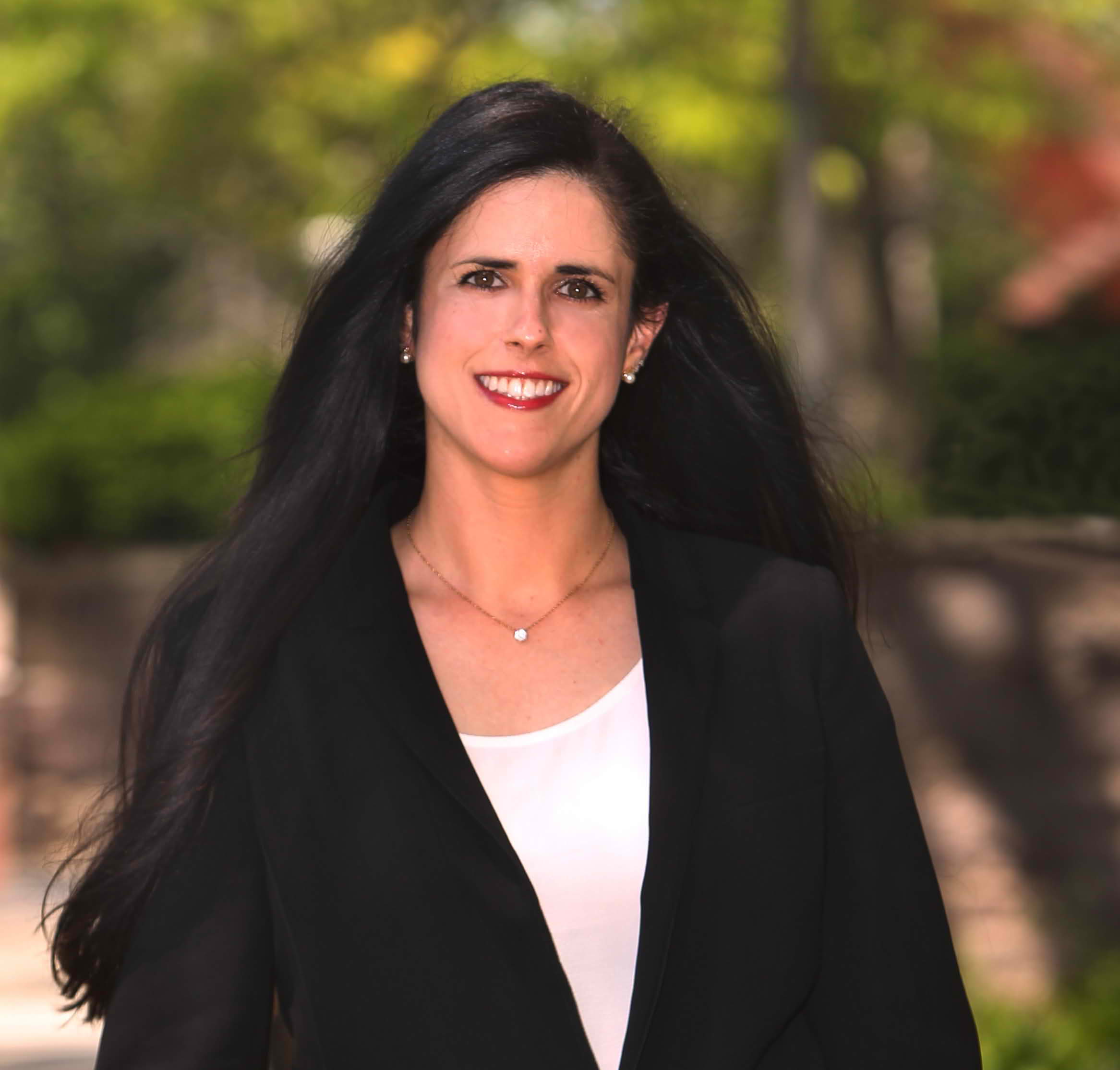 A woman with long dark hair, wearing a black blazer over a white top, smiles outdoors with sunlight filtering through green trees—a vibrant scene that reflects the spirit of sustainability at Cority.