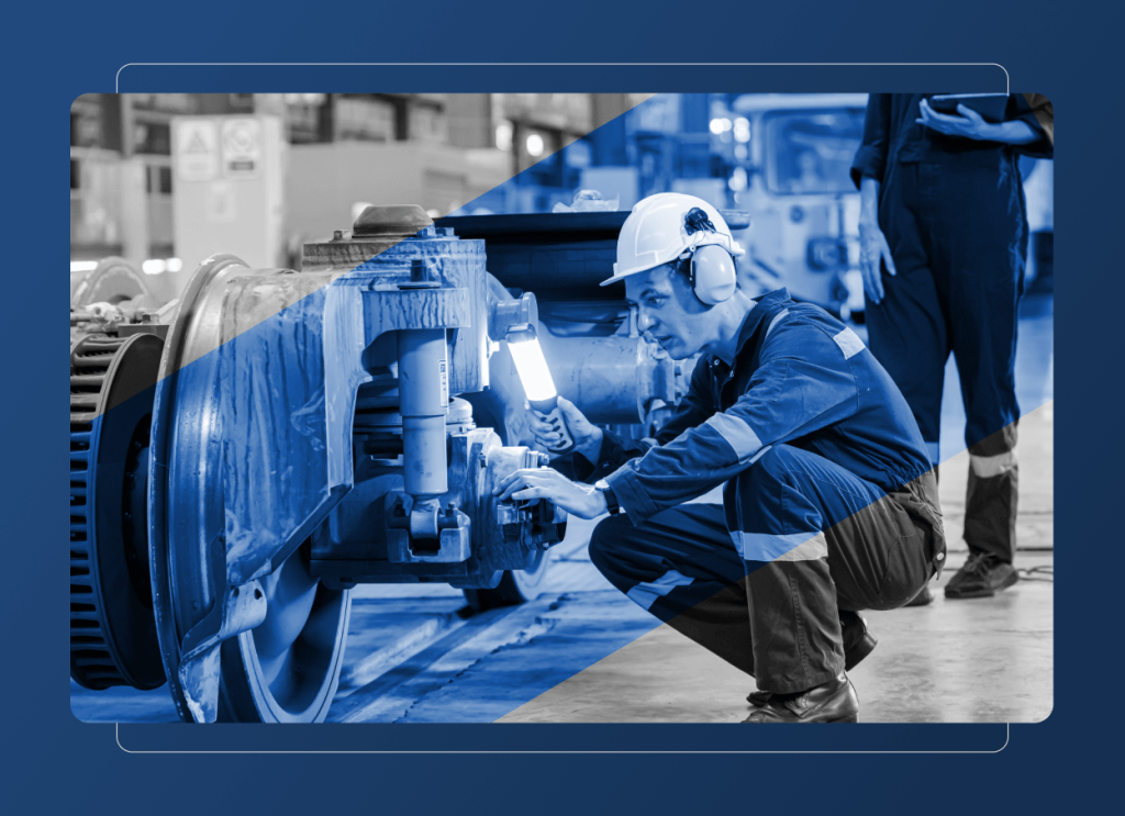 A worker wearing a hard hat and ear protection inspects a train wheel with a flashlight in a manufacturing industry setting, while another person stands nearby holding a tablet.