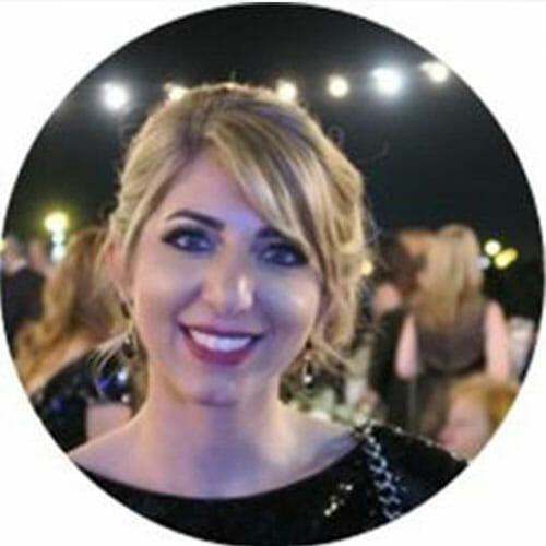 A woman with blonde hair and red lipstick smiles at the camera during an outdoor nighttime event celebrating sustainability at Cority, with blurred people and string lights glowing in the background.