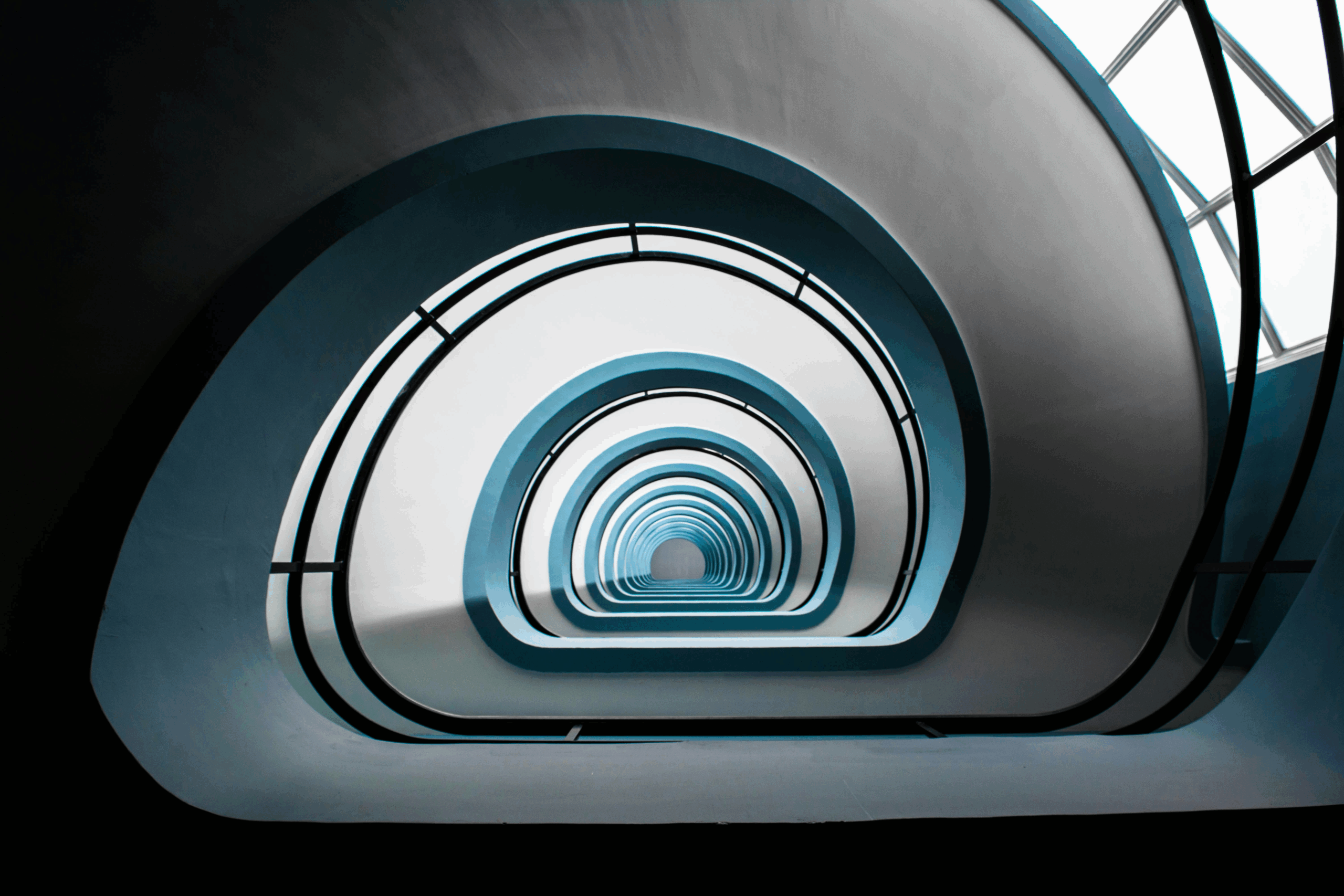 A view looking up through a spiral staircase with smooth white and blue curves, resembling the streamlined effect of ehs software consolidation, creating a symmetrical, tunnel-like pattern that leads the eye toward a bright light at the top.