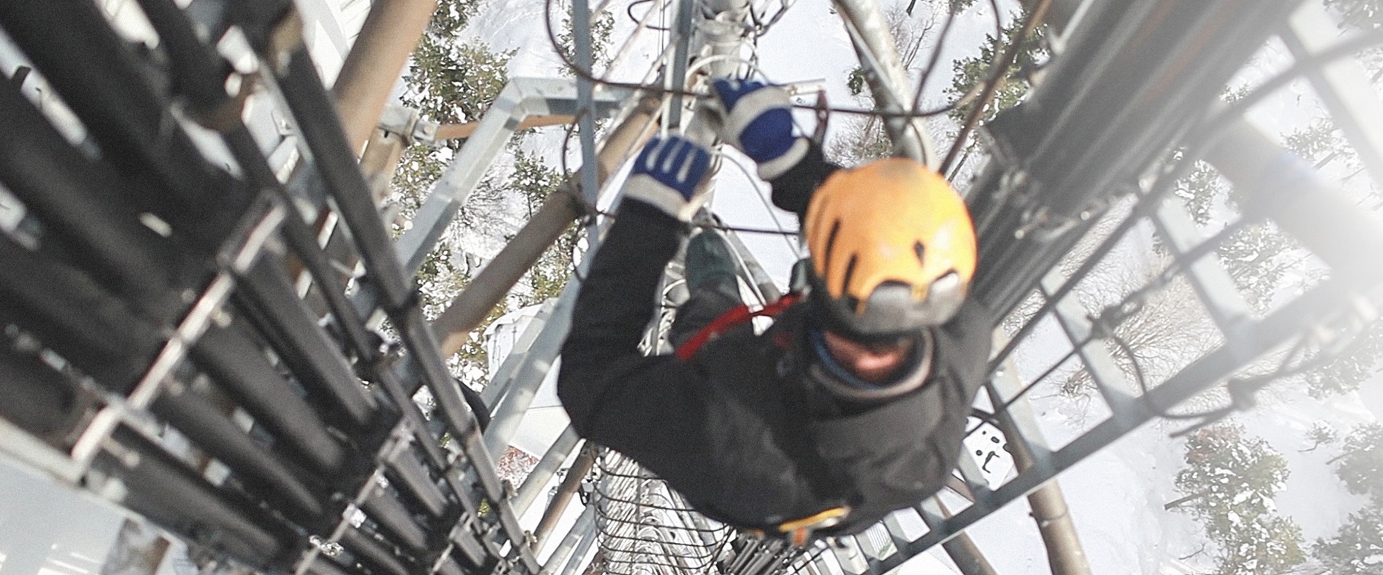 A worker wearing a yellow helmet and safety gear climbs a tall communications tower as part of digital incident management, gripping metal rungs beside large cables, with a snowy landscape visible below.