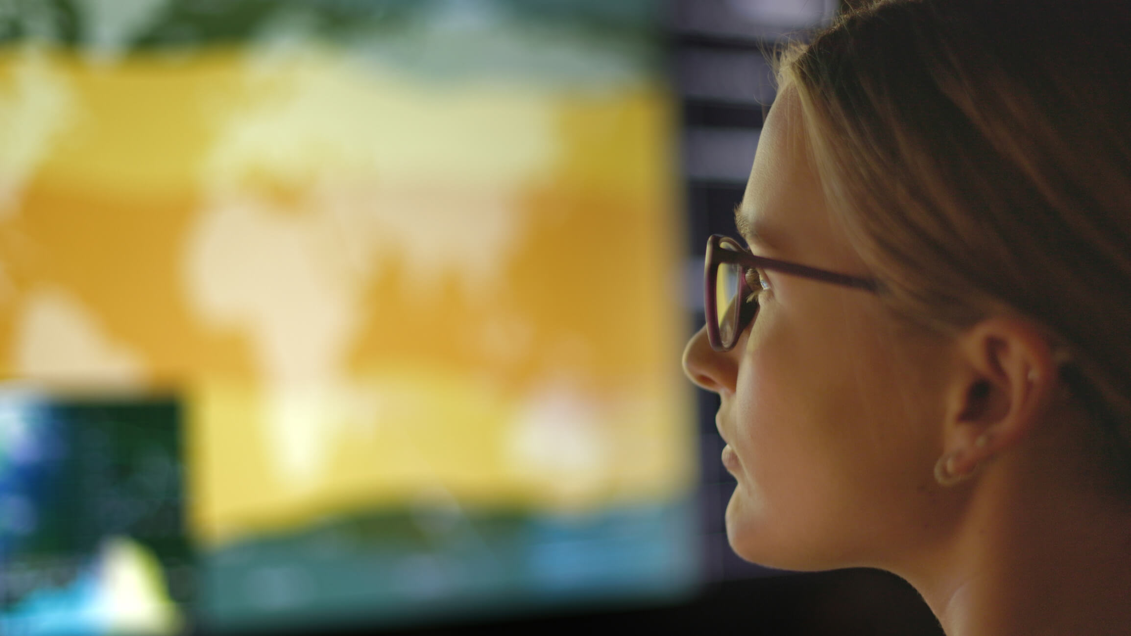 A woman wearing glasses looks at a large, blurred screen displaying a yellow-toned world map, suggesting data analysis or monitoring of leading safety indicators in a technology-driven environment.