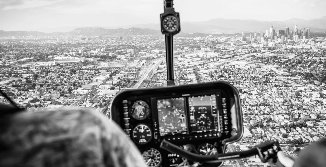 Black and white view from inside a helicopter cockpit flying over a sprawling city, with instrument panels visible and buildings, roads, and mountains in the distance.