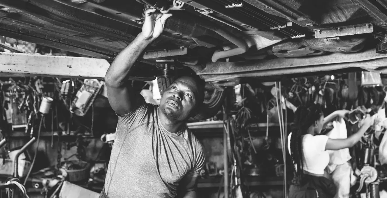 A mechanic inspects the underside of a raised vehicle in a garage, with tools and car parts visible in the background. Another person works at a bench. The image is in black and white.